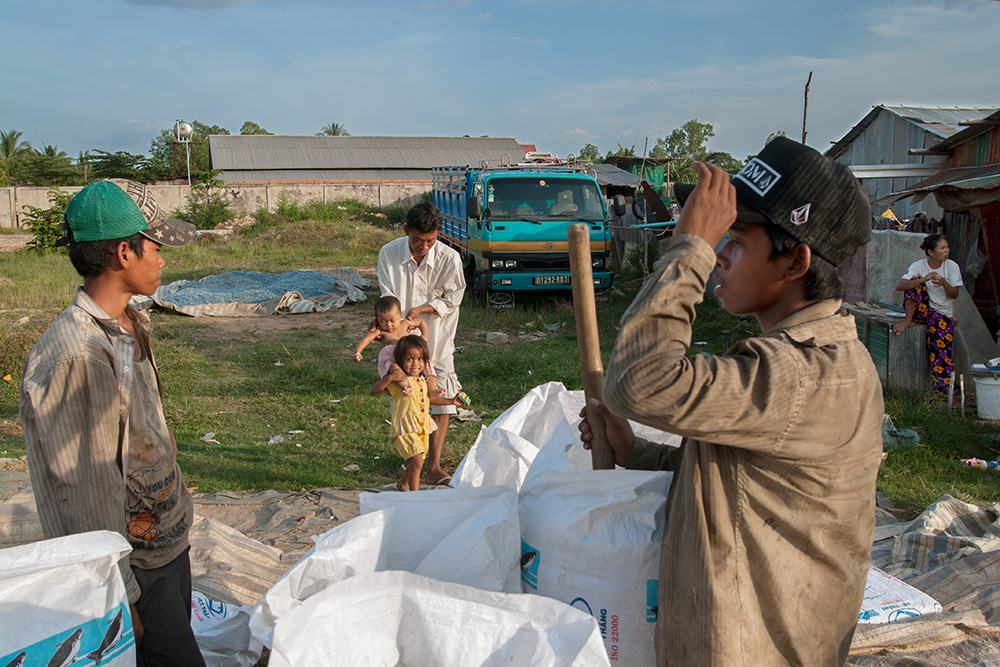 Young men load minced recycled plastic into bags for sale. A warehouse owner plays with his kids at the background.